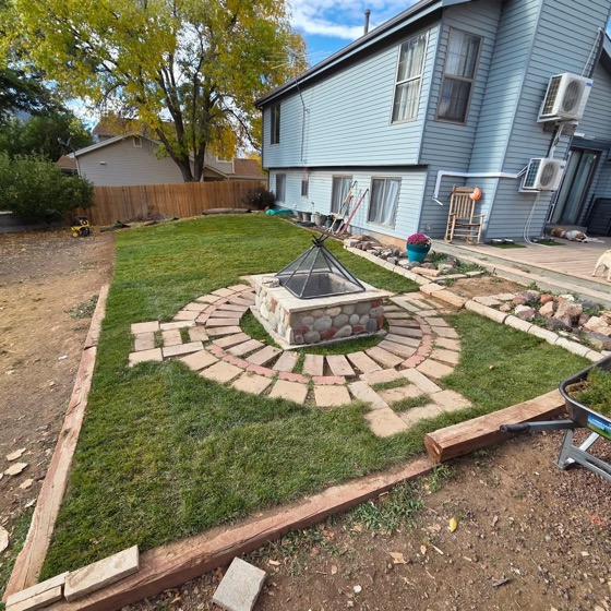 A finished lawn with a stone fire pit inside a ring of brick pavers, behind a blue-gray house
