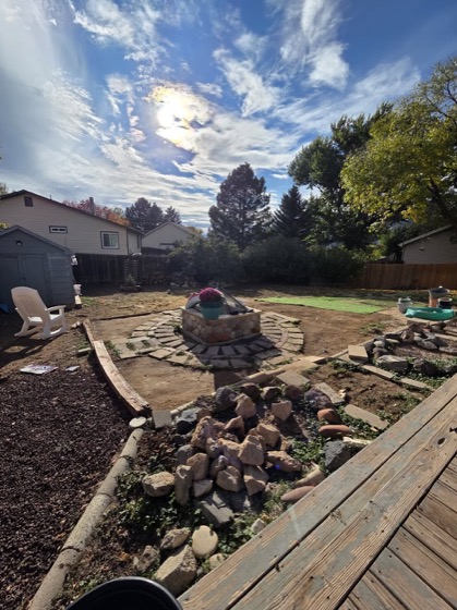 A backyard mid-project in afternoon sun, with a deck in the foreground and a stone fire pit ahead